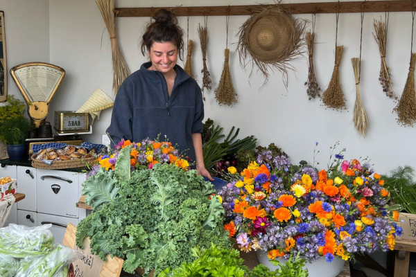 Smiling woman stands behind a market table, arranging vibrant bouquets of orange, yellow, purple, and pink flowers beside piles of curly kale and leafy greens, with hanging dried herbs, a straw hat, and an old scale on shelves behind her.