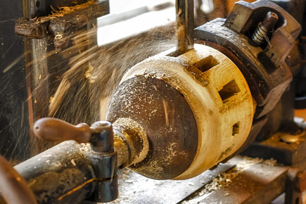 Woodworking lathe holds a rotating log while a drill bit bores into its center, spraying wood shavings outward. The cylindrical blank shows square cutouts, and the workshop machine bed is dusted with sawdust.