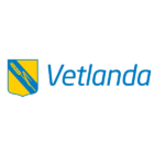 Vetlanda kommuns logotyp visas: gul och blå vapensköld med diagonala blå band och ett veteax, följt av ordet ”Vetlanda” i blå kursiv text på svart bakgrund.