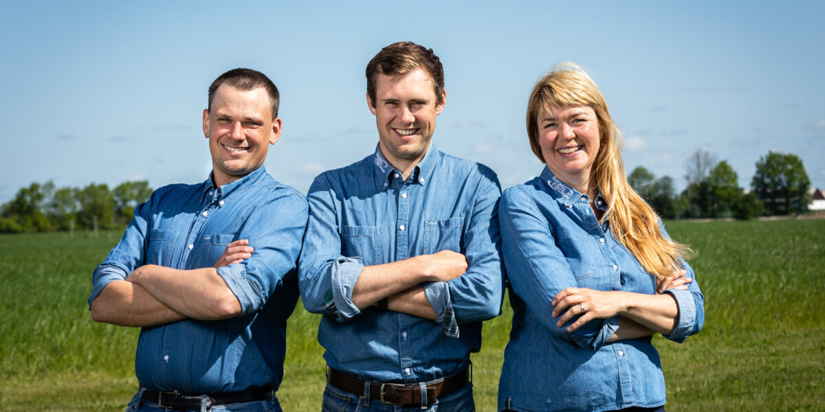 Tre personer står tätt ihop med armarna i kors och ler mot kameran, alla i blå denimskjortor. De är fotograferade utomhus på en grön åker med blå himmel och träd i bakgrunden.