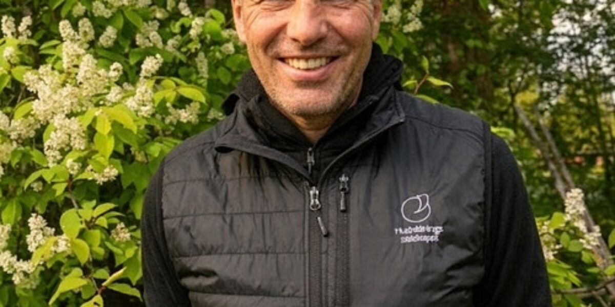 Smiling man stands outdoors with hands behind his back, wearing a black quilted vest with a small white logo, framed by lush green foliage and clusters of white blossoms in warm daylight.