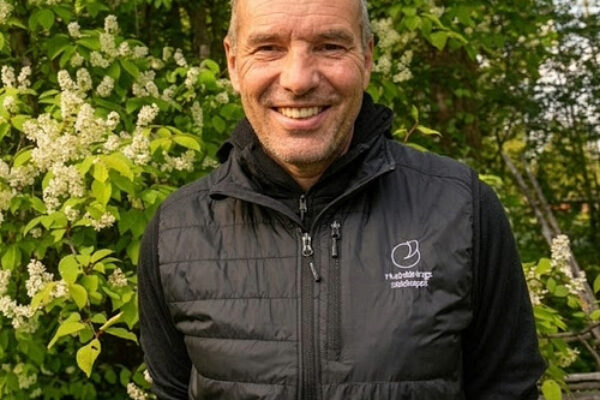 Smiling man stands outdoors with hands behind his back, wearing a black quilted vest with a small white logo, framed by lush green foliage and clusters of white blossoms in warm daylight.