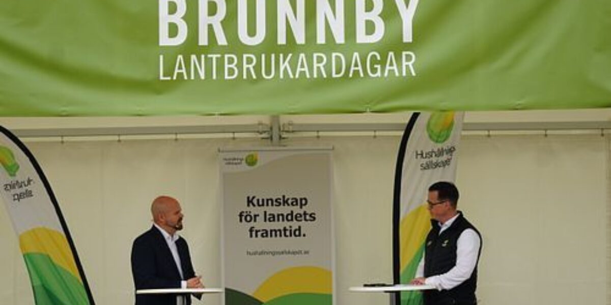Två män står på en scen och samtalar vid var sitt ståbord inför en publik, under ett grönt tält märkt ”BRUNNBY LANTBRUKARDAGAR”, med banderolltexten ”Kunskap för landets framtid.” och ”Hushållningssällskapet”.