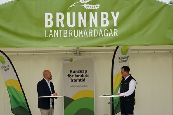 Två män står på en scen och samtalar vid var sitt ståbord inför en publik, under ett grönt tält märkt ”BRUNNBY LANTBRUKARDAGAR”, med banderolltexten ”Kunskap för landets framtid.” och ”Hushållningssällskapet”.