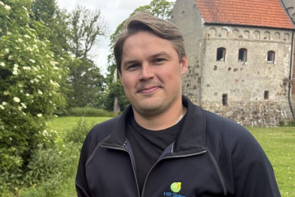 Man i mörk jacka står och ler mot kameran på en grön gräsmatta. På bröstet syns texten ”HIR Skåne”. I bakgrunden finns blommande buskar och en gammal stenbyggnad med rött tegeltak.