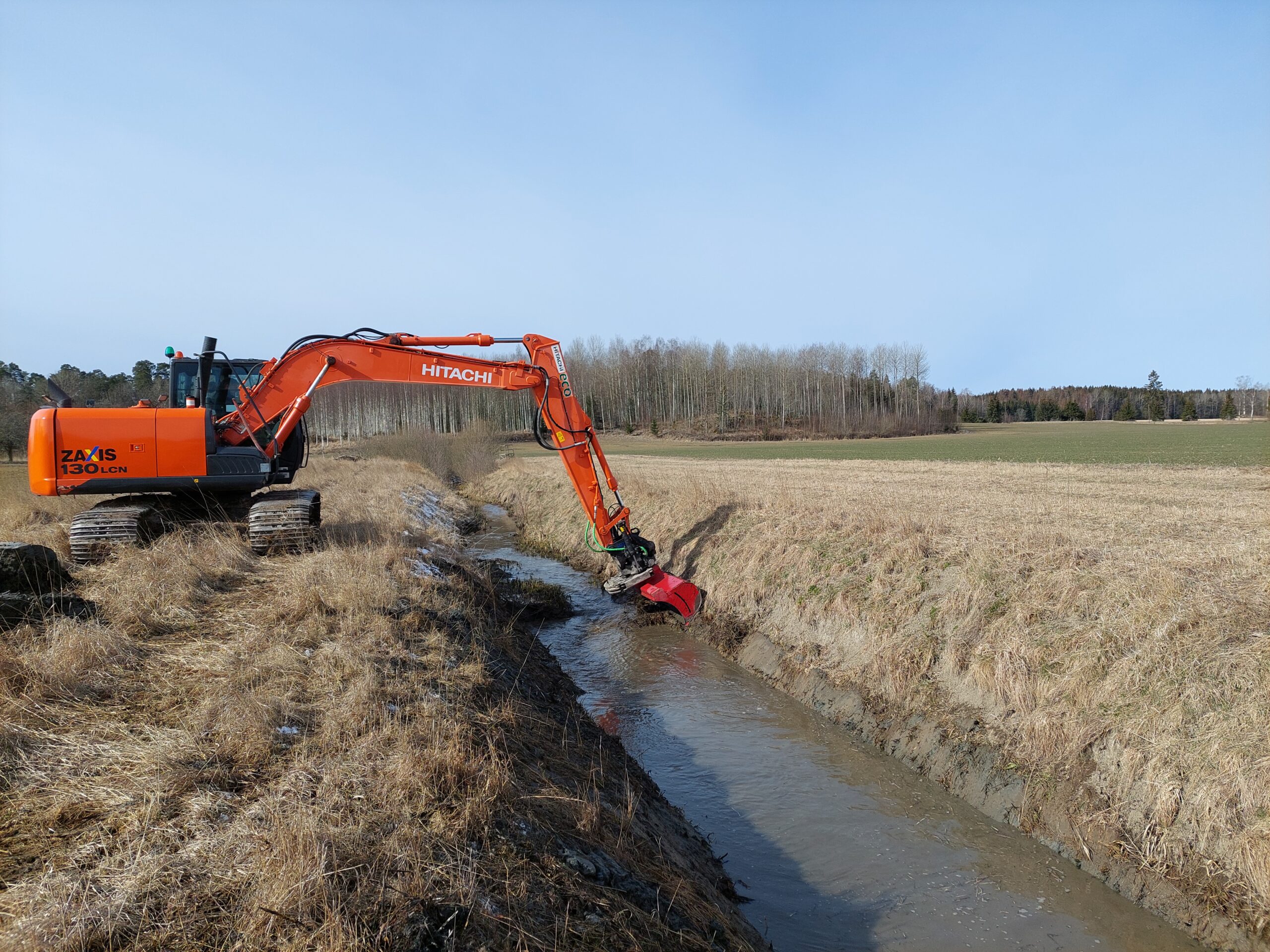 Orange bandgrävare från Hitachi märkt “HITACHI” och “ZAXIS 130 LCN” rensar ett dike med röd klipp- eller slaghack, bredvid ett lerigt vattendrag på åkermark med skog i bakgrunden.