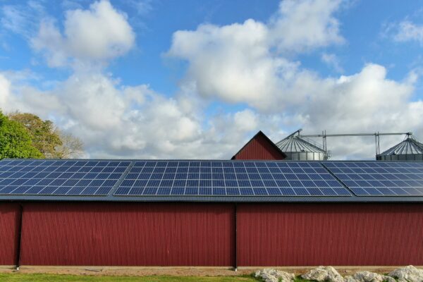 Solar panels cover the roof of a long red barn, generating power on a rural farm. Two metal grain silos and a small gable rise behind the roof under a bright, cloudy sky.