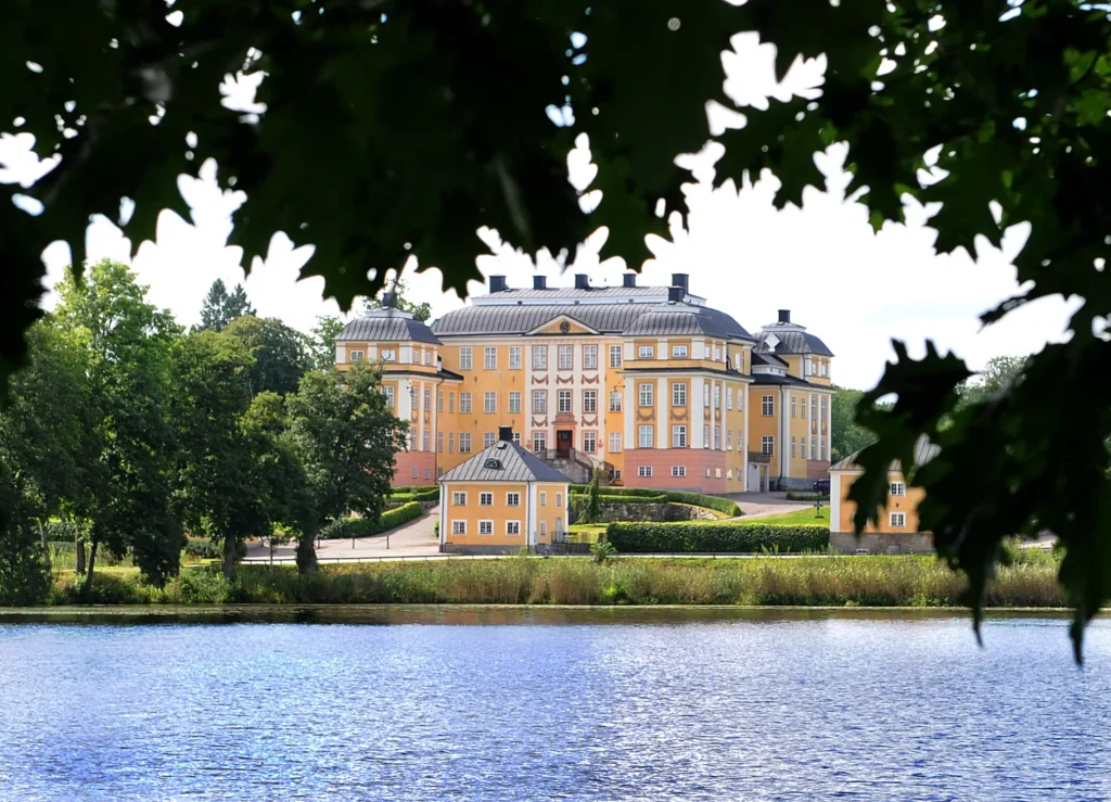 Gul slottliknande byggnad med grått tak står vid en sjö och omges av grönskande träd och välklippta häckar. Fotot är taget på avstånd och ramas in av mörka löv i förgrunden.