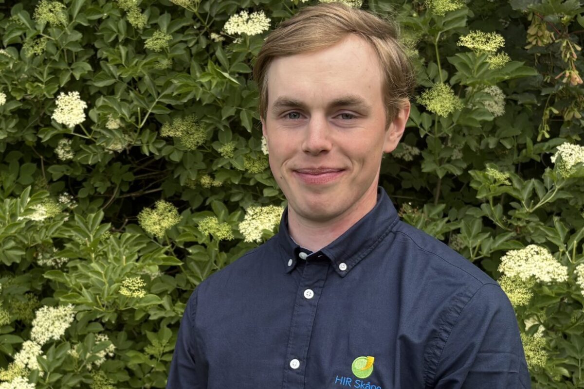 Leende man står utomhus framför en tät grön buske med många små vita blomklasar. Han bär mörkblå skjorta med broderad logotyp och texten “HIR Skåne” på bröstet.