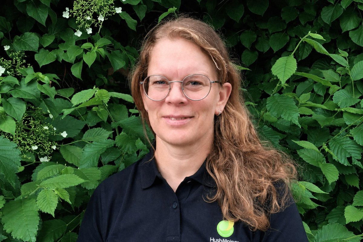 Woman with long light-brown hair and round glasses faces the camera with a slight smile, standing in front of dense green foliage with small white blossoms, wearing a black polo shirt with a “Hushållningssällskapet” logo.