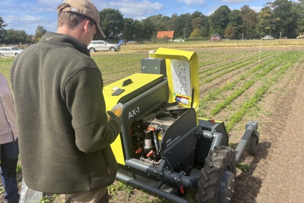 Man in a cap inspects an open yellow farm machine labeled “AX-1,” looking at its controls and exposed engine beside neat crop rows in a field, with cars, trees, and a small red-roofed building in the background.