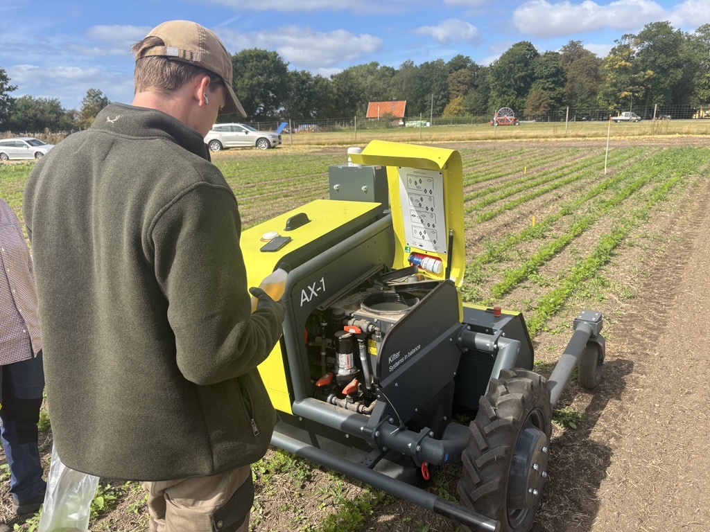 Man in a cap inspects an open yellow farm machine labeled “AX-1,” looking at its controls and exposed engine beside neat crop rows in a field, with cars, trees, and a small red-roofed building in the background.