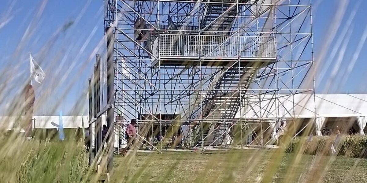Hög ställningsbyggd läktare med trappor och plattformar står på en gräsplan, med personer som rör sig vid basen. Överst hänger en banderoll: “KUNSKAP FÖR LANDETS FRAMTID”, “www.hushallningssallskapet.se”, “Hushållnings sällskapet”.