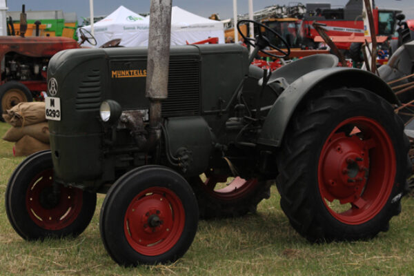 Grön vintage-traktor av märket Munktells står parkerad på gräs med stora bakhjul och röda fälgar. Nummerplåt “H 6293” syns framtill. I bakgrunden finns andra traktorer, tält och besökare på utställning.