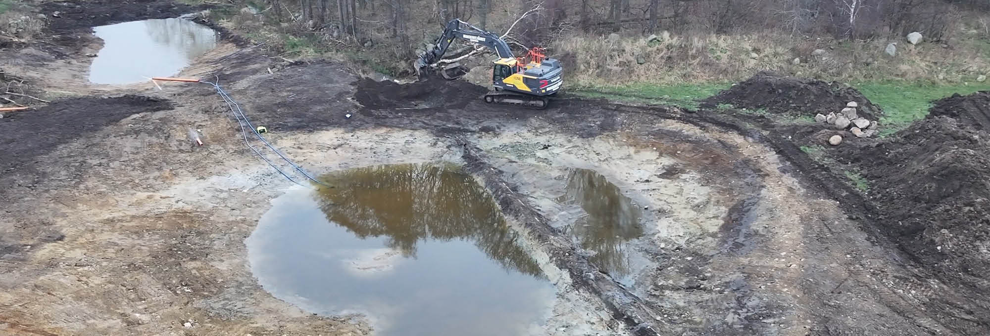 Bandgrävmaskin gräver och flyttar mörk jord på en lerig arbetsplats vid en vattenfylld grop, med slangar över marken och jord- och stenmassor längs kanten inom LEVA Halland.