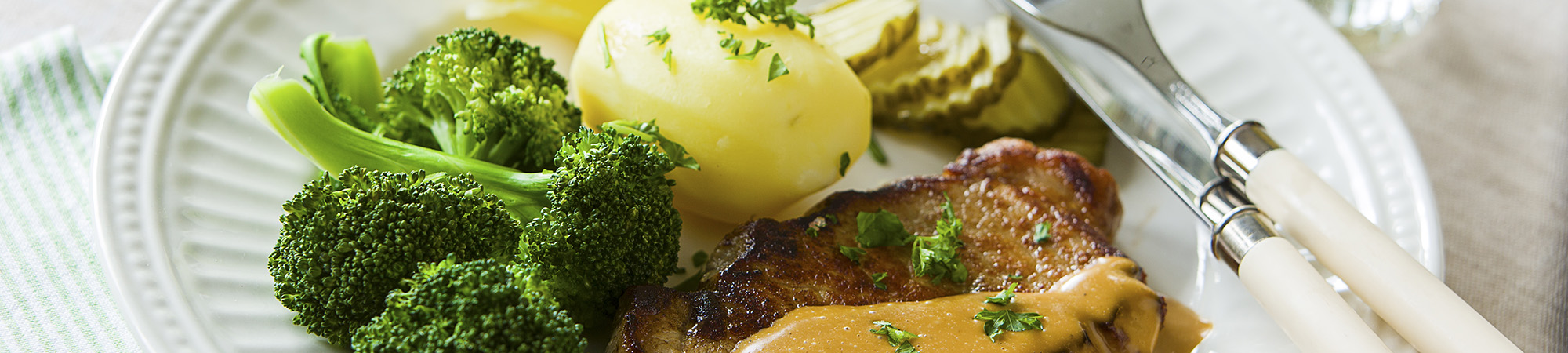 Tallrik med middag serveras med stekt kött med gräddig sås och persilja, kokt potatis, broccoli och skivade pickles. Bestick med vita skaft ligger bredvid på en ljus duk, som headerbild för matwebbinar.