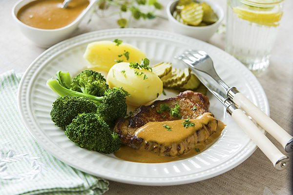 Tallrik med köttbit i brun sås serveras med kokt potatis och broccoli. Skivad inlagd gurka ligger bredvid, och bestick vilar på kanten. I bakgrunden syns soppskål och glas med citronvatten.