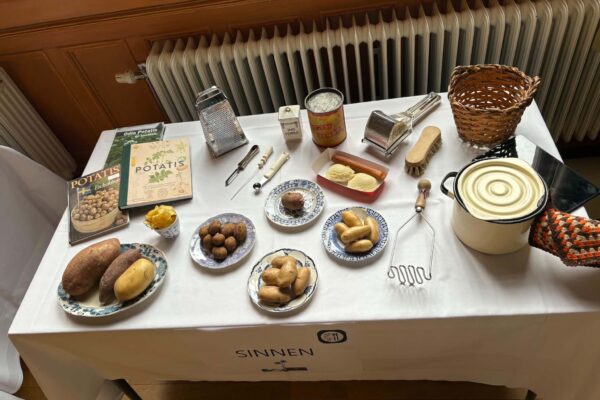 Table display presents potatoes and preparation tools, arranged like a small exhibit: plates of different potato varieties, cookbooks titled “POTATIS,” graters, peeler, potato masher, pot, basket, and seasonings labeled “HVIT PEPPAR.” Text on table front: “SINNEN.”