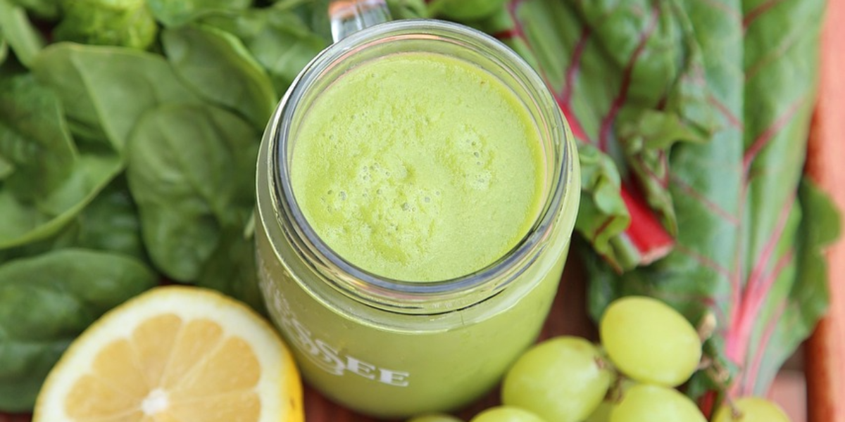 Glasburk med grön smoothie står på ett träbord, omgiven av färska bladgrönsaker. Bredvid ligger halverad grapefrukt och citron samt en klase gröna druvor, vilket ger en känsla av hälsosam, fräsch ingrediensmix.