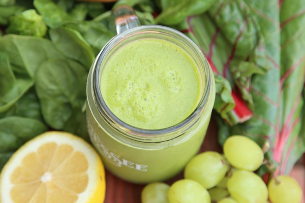 Glasburk med grön smoothie står på ett träbord, omgiven av färska bladgrönsaker. Bredvid ligger halverad grapefrukt och citron samt en klase gröna druvor, vilket ger en känsla av hälsosam, fräsch ingrediensmix.