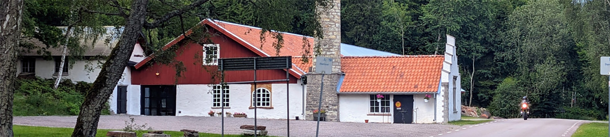 Gammal vit och röd byggnad med rött tegeltak och hög stenmurad skorsten står vid en landsväg; en motorcyklist kör förbi på vägen, med skog och träd som bakgrund.