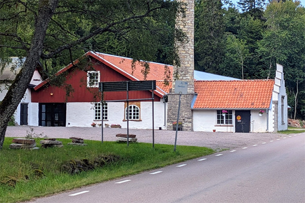 Röda och vita byggnader med tegeltak står intill en landsväg, med ett högt stenmonument eller skorsten i mitten. En vägskylt syns framför och skog omger området i bakgrunden.