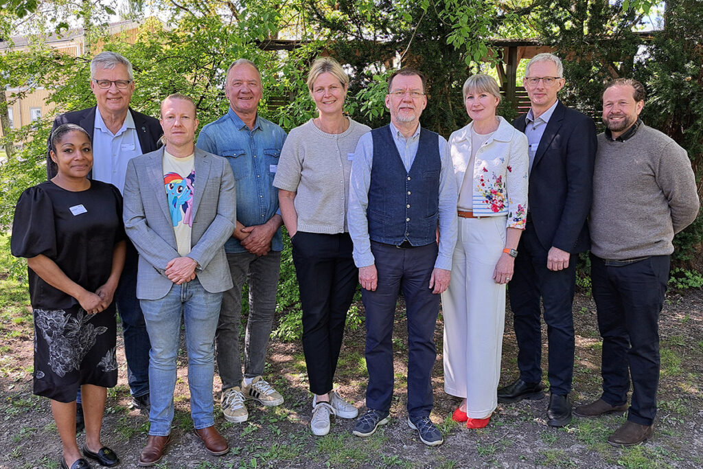 Åtta personer står uppställda och poserar för ett gruppfoto utomhus. De bär namnskyltar och är klädda i blandade kavajer, skjortor och vardagskläder, med grönska och träd i bakgrunden.