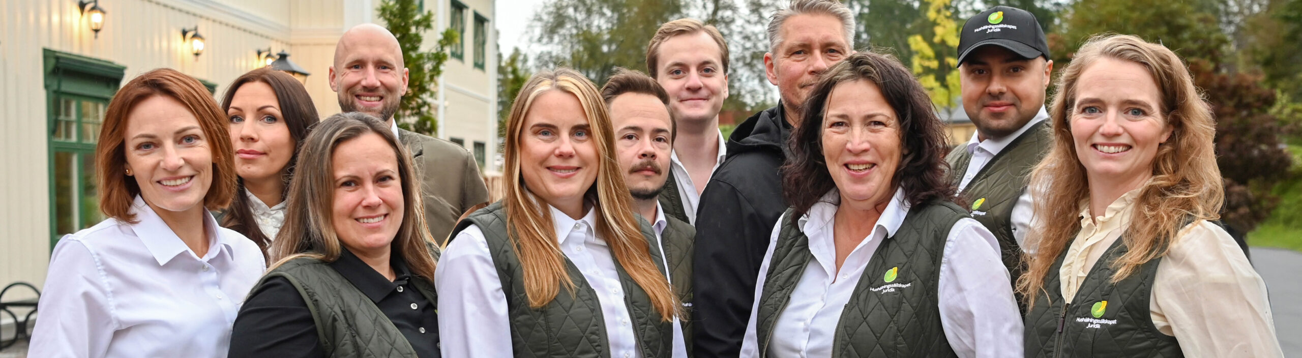 Grupp på tio personer står tätt tillsammans och ler mot kameran utomhus vid en ljus byggnad och träd. Flera bär gröna västar och keps med loggan och texten ”Nutrilettkapet Järla”.