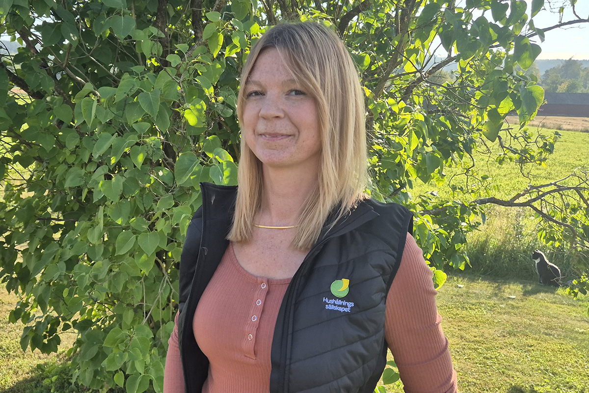 Kvinna med axellångt blont hår står och ler lätt framför ett lövigt träd i solsken. Hon bär svart väst med texten ”Hushållningssällskapet” över en rostfärgad tröja, med åker och gård i bakgrunden.