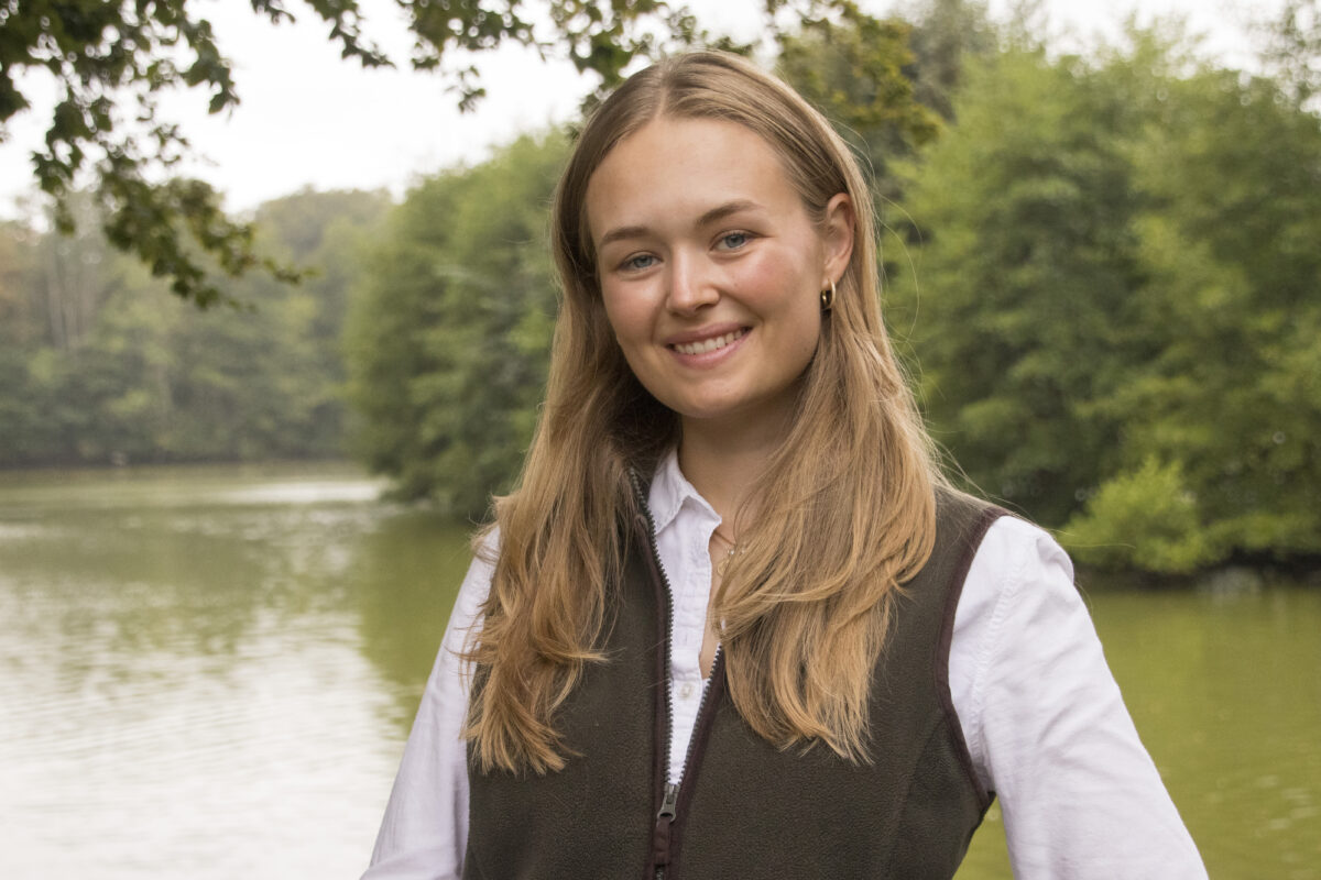 Leende ung kvinna med långt blont hår ser in i kameran och står vid en lugn å med skog i bakgrunden. Hon bär vit skjorta och mörk väst, med grenar som ramar in överkanten.