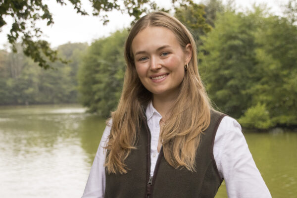 Leende ung kvinna med långt blont hår ser in i kameran och står vid en lugn å med skog i bakgrunden. Hon bär vit skjorta och mörk väst, med grenar som ramar in överkanten.