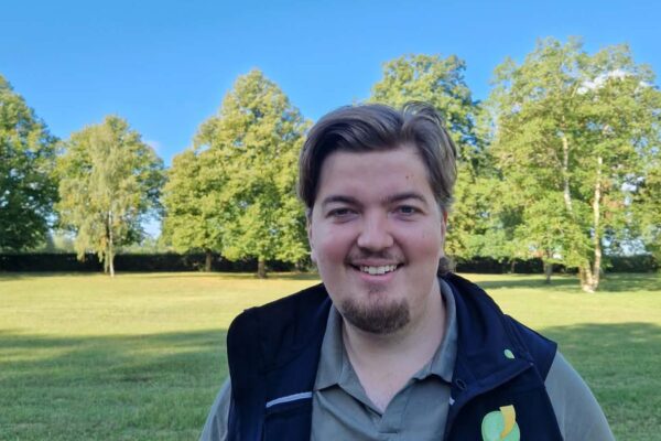 En leende man med kort skägg tittar in i kameran och står utomhus i en park. Han bär grön pikétröja och mörk väst med en rund grön logga, med träd och blå himmel i bakgrunden.
