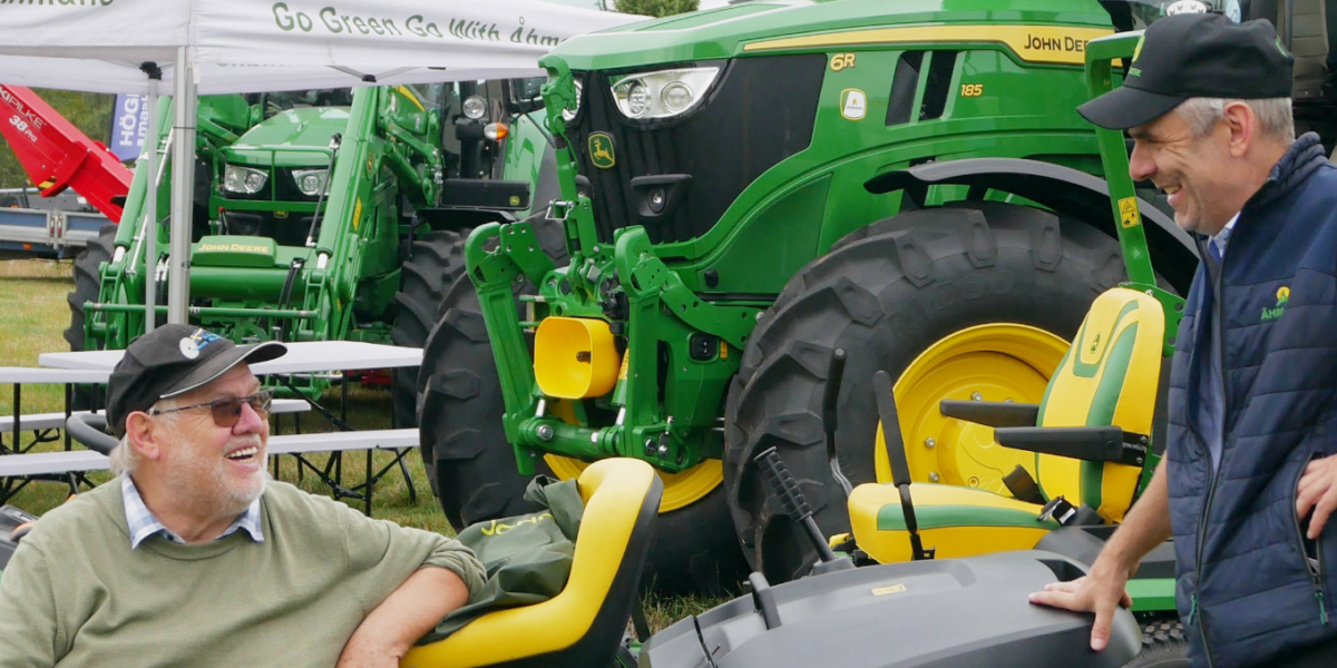 Två män pratar och skrattar vid en gulgrön John Deere gräsklippare på en utomhusmässa. I bakgrunden syns stora gröna John Deere-traktorer, tält och bänkbord samt texten “Go Green Go With John Deere”.