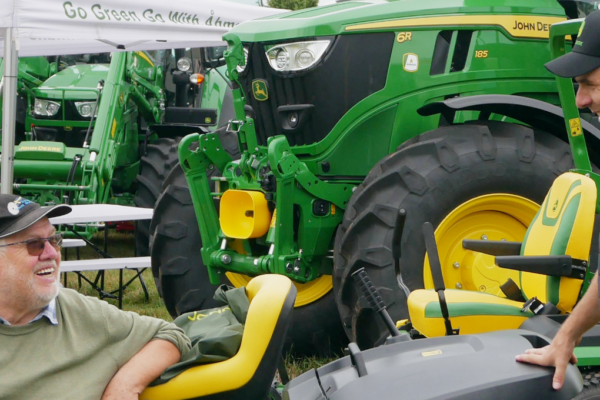Två män pratar och skrattar vid en gulgrön John Deere gräsklippare på en utomhusmässa. I bakgrunden syns stora gröna John Deere-traktorer, tält och bänkbord samt texten “Go Green Go With John Deere”.