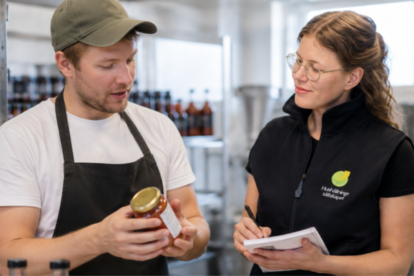 Man i keps och förkläde visar en burk med innehållsetikett och pratar med kvinna som antecknar. De står i en livsmedelslokal med hyllor och flaskor i bakgrunden. Text på väst: ”Hushållningssällskapet”.