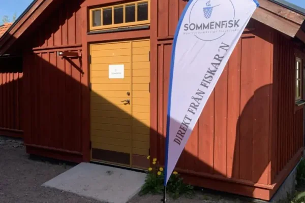 Röd träbyggnad med gul dörr står i soligt väder, och en vit reklamflagga framför entrén vajar lätt. Text på flaggan: ”SOMMENFISK” och ”DIREKT FRÅN FISKAR’N”. Grusplan och klarblå himmel runtom.