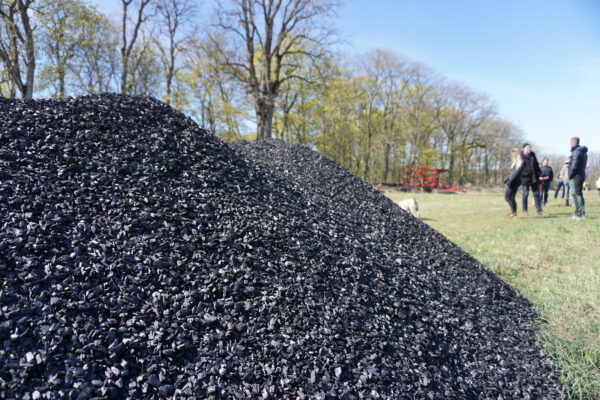 Stor hög av svart biokol ligger i förgrunden på en gräsplan, medan en liten grupp människor står och samtalar i bakgrunden vid träd och jordbruksutrustning under klar blå himmel.