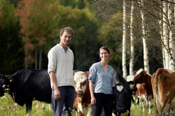 En man och en kvinna står leende i en grön hage bland betande kor. Runt dem syns svartvita och brunvita kor, med björkar och skog i bakgrunden, en lugn gårdsmiljö.