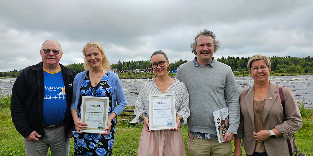 Fem personer poserar och ler vid en älv med skog och molnig himmel i bakgrunden; två i mitten håller inramade diplom med texten ”DIPLOM”, och mannen bredvid håller en broschyr.