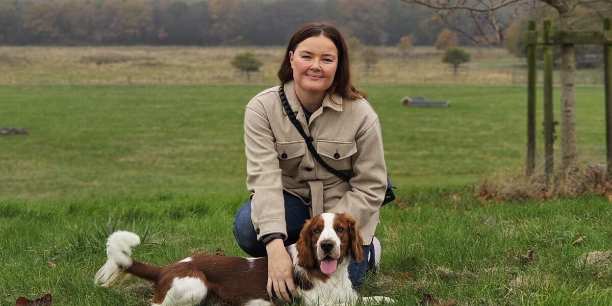 Kvinna i beige jacka sitter på huk i gräset och klappar en brunvit hund som ligger framför henne med tungan ute. I bakgrunden syns öppna fält, höstfärgade träd och en mulen himmel.