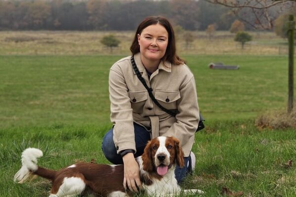 Kvinna i beige jacka sitter på huk i gräset och klappar en brunvit hund som ligger framför henne med tungan ute. I bakgrunden syns öppna fält, höstfärgade träd och en mulen himmel.