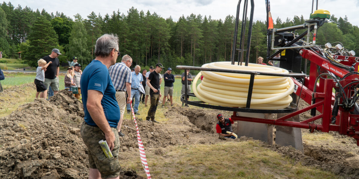 Åskådare står vid en uppgrävd ränna och tittar när en röd maskin med stor gul slangrulle lägger ner slang i marken, medan en arbetare sitter i schaktet; skog och åker i bakgrunden. Text: “www.lasole”.