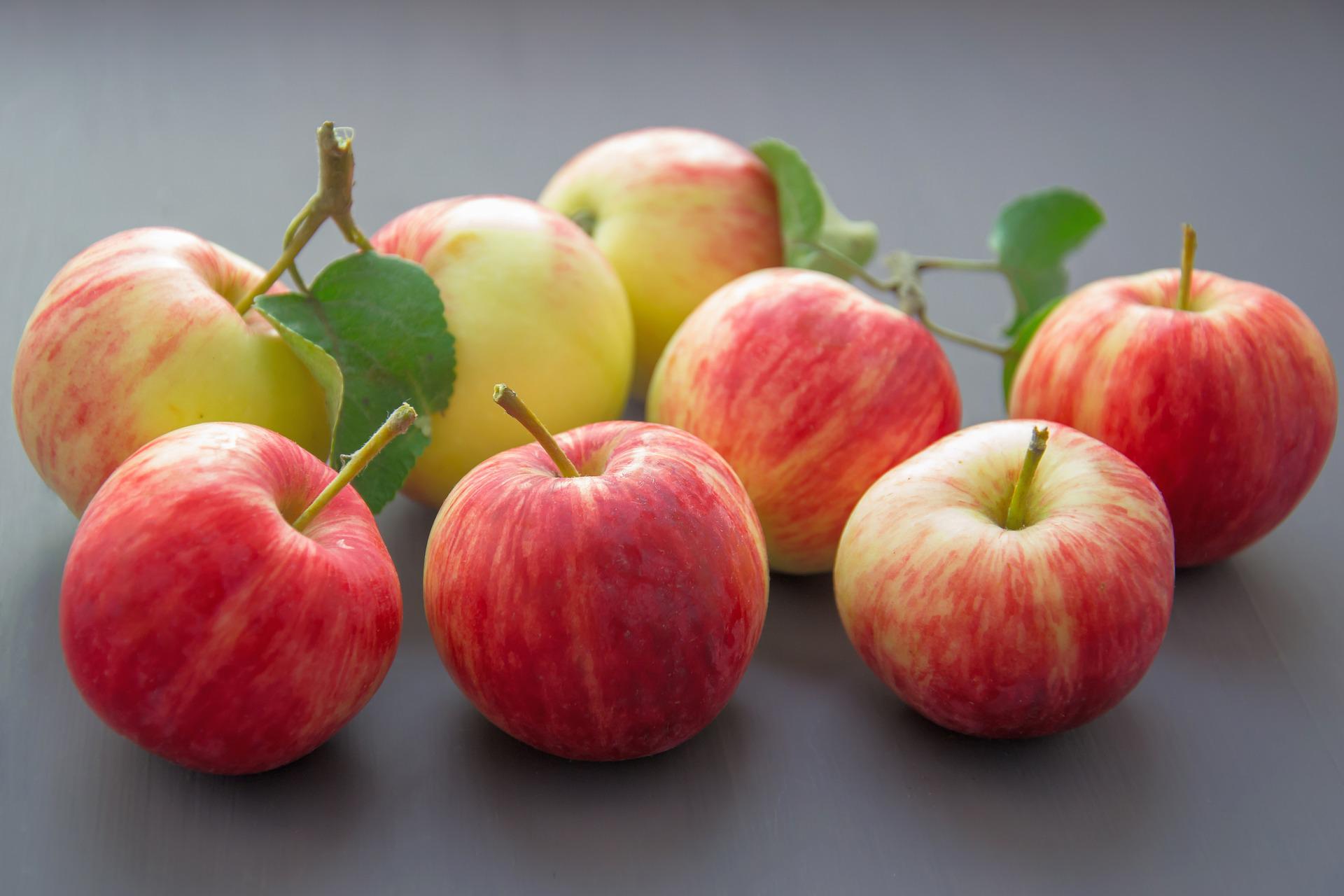 Sex rödrandiga äpplen ligger tätt samlade på en mörk bordsyta, några med stjälkar och gröna blad kvar. Närbild med mjuk grå bakgrund framhäver frukternas blanka skal och färgskiftningar.