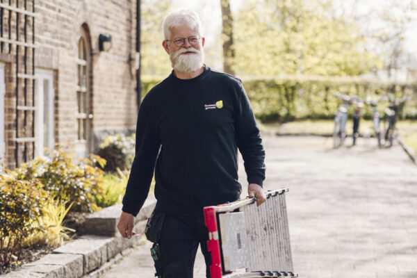 Äldre vaktmästare med vitt skägg går på en trottoar och bär en ihopfälld stege. Han har arbetskläder med texten ”Onestads gymnasium” och passerar en tegelbyggnad i soligt väder.