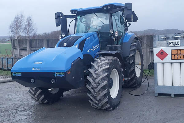 Blå traktor står parkerad på en gårdsplan och tankas via en slang vid en station för biogas. På stationen syns skyltar med ”FYLD” och siffrorna ”23 1971”.
