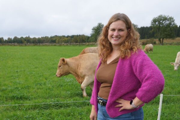 Leende kvinna med lockigt hår står med handen på höften vid ett elstängsel, klädd i rosa kofta och jeans. Bakom henne betar ljusbruna kor på en grön äng med träd i bakgrunden.