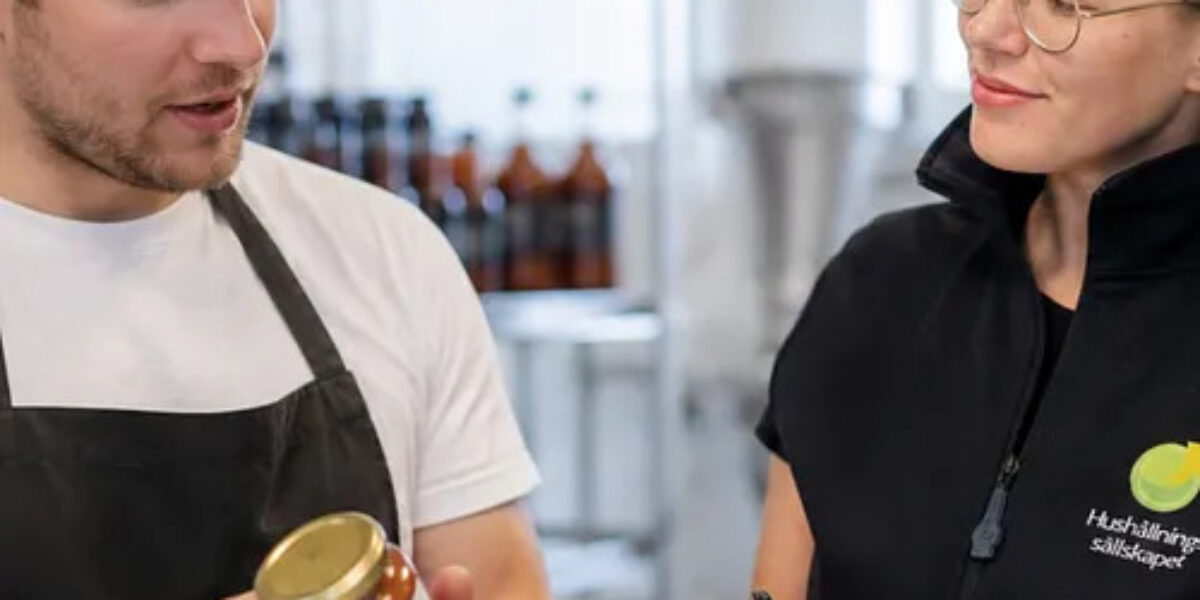 En man i keps och förkläde håller upp en glasburk med produktetikett och förklarar, medan en kvinna i glasögon och väst med texten ”Hushållningssällskapet” antecknar i ett block i en livsmedelslokal.