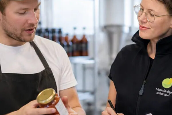 En man i keps och förkläde håller upp en glasburk med produktetikett och förklarar, medan en kvinna i glasögon och väst med texten ”Hushållningssällskapet” antecknar i ett block i en livsmedelslokal.