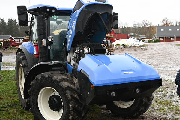 Blå traktor står på en lerig gårdsplan med motorhuven uppfälld, så att motorrummet syns. I bakgrunden finns gårdsbyggnader, maskiner och snöhögar, vilket ger intryck av service eller demonstration utomhus.