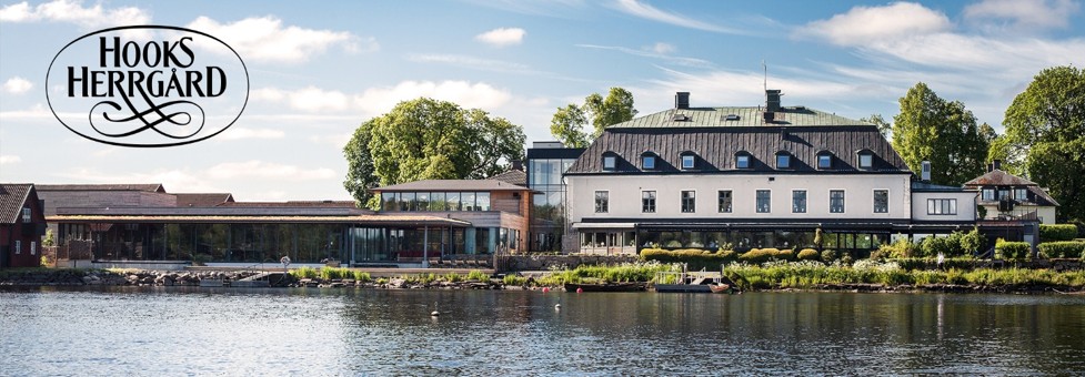 Hooks Herrgård syns på andra sidan en stilla sjö, med huvudbyggnad och en modern glasdel längs strandkanten. Grönska och sommarhimmel ramar in anläggningen. Text i logga uppe till vänster: “HOOKS HERRGÅRD”.